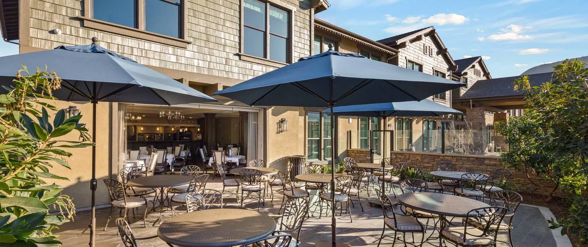 Outdoor dining patio at Oakmont of Agoura Hills featuring elegant metal tables with umbrellas, cushioned chairs, and a view of the community’s modern building under clear blue sky.