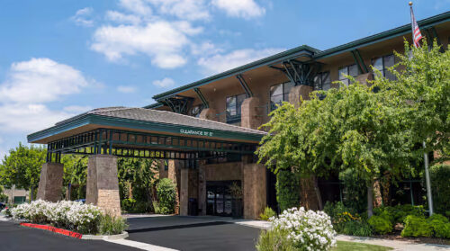 Front exterior of Hampton Inn & Suites Agoura Hills featuring covered entrance with stone pillars, green trim, landscaped trees, and a clear blue sky.