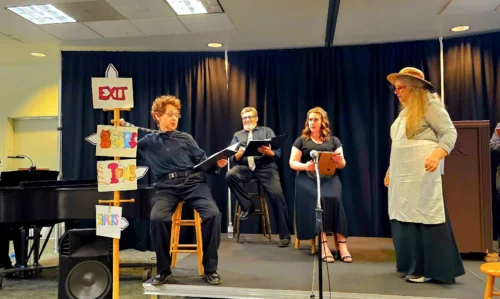 Actors performing a stage reading at Hillcrest Center for the Arts in Thousand Oaks, with a piano and colorful handmade signs on stage.