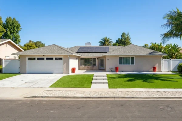 A wide-angle, eye-level shot of a single-story ranch-style residential home under a clear blue sky. Bright, direct sunlight illuminates the beige exterior, white garage door, and solar panels installed on the grey shingled roof. A perfectly manicured green lawn is split by a concrete walkway leading to the entrance, where the house number "143" is visible. The composition is balanced with palm trees and white fencing on the sides, creating a clean, suburban California aesthetic.