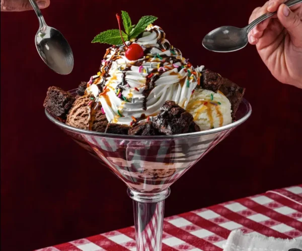 Brownie sundae in martini glass with whipped cream, ice cream scoops, sprinkles, and cherry, two spoons reaching in.