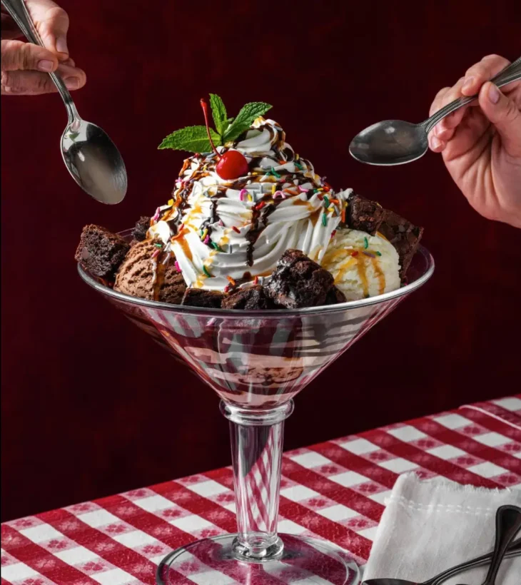 buca-di-beppo-brownie-sundae-dessert Brownie sundae in martini glass with whipped cream, ice cream scoops, sprinkles, and cherry, two spoons reaching in.