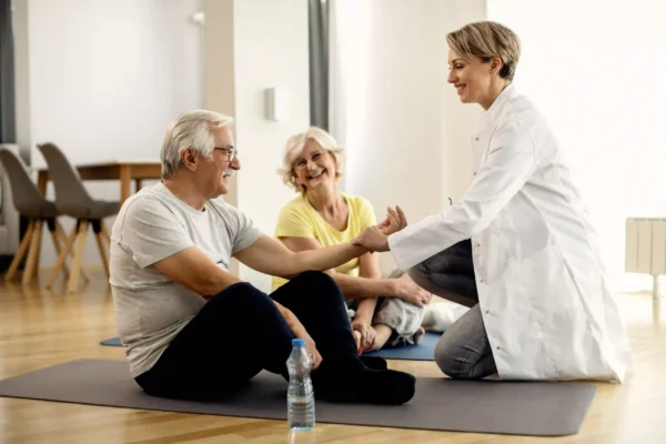 House-call doctor checking an older man’s pulse while his partner smiles during a wellness visit at an assisted living home