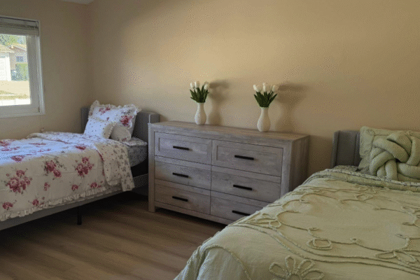 Shared bedroom at Anita’s Care Villa with two beds, a dresser, and natural light from a window.