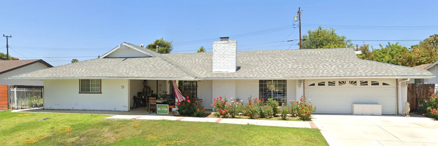 Exterior view of RCFE Arianah Place, a white single-story residential care facility for the elderly at 53 Wales Street, Thousand Oaks, featuring a manicured lawn, rose bushes, and a classic suburban ranch-style architecture under a clear blue sky.