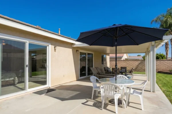 Sunny backyard patio of a single-story home with glass doors, round table, white chairs, and large dark umbrella.