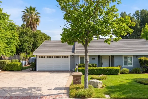 A perfectly centered, straight-on frontal view of a single-story house. The composition is entirely flat with no perspective distortion, captured at exact eye level. Bright, warm natural sunlight illuminates the scene, creating high contrast, vibrant color richness, and soft, realistic shadows across the fresh green lawn and patterned driveway. The lighting gives the image a clean, crisp, and highly polished professional real estate photography aesthetic. A small house number is faintly visible on the wall right next to the front door under the porch.