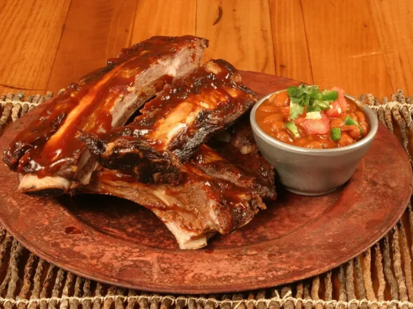 Plate of barbecue ribs glazed with sauce, served with a side of baked beans on a rustic wooden table.