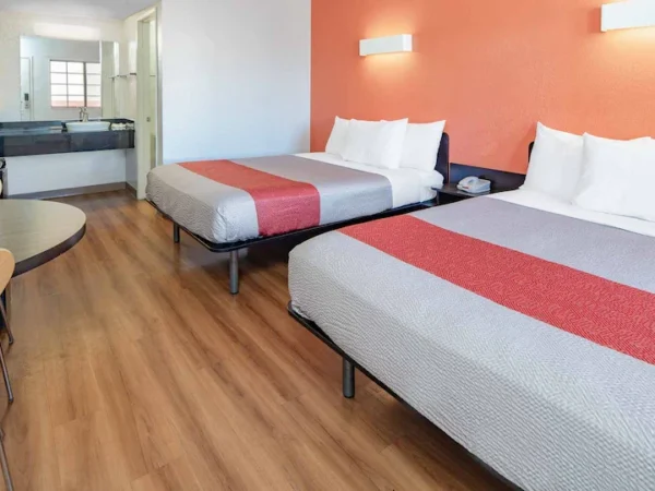 Motel room with two beds, wood-look flooring, small table, and open vanity area.