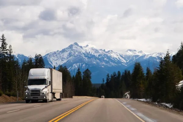 Semi-truck driving on a highway with snow-capped mountains in the background.