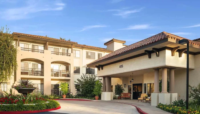 A clear, sun-drenched architectural shot of the Atria Hillcrest entrance under a bright blue sky. The Mediterranean-style building features light stucco walls, dark wooden accents, and a classic red-tile roof. The number "405" is clearly visible in black centered on the white portico above the driveway. The foreground showcases a manicured circular driveway with a lush, vibrant flowerbed of red blooms and green shrubbery.
