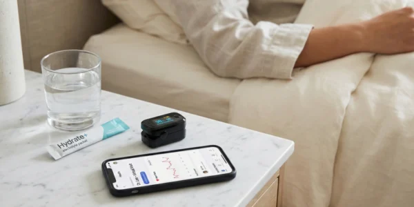 A close-up, high-angle shot of a white marble nightstand in a sunlit room. On the table sits a clear glass of water, a white and teal sachet labeled "Hydrate+ electrolyte packet," and a black digital pulse oximeter showing a "98%" reading. A smartphone lies flat, displaying a medical app with a heart rate graph and the text "Dr. Lee - Virtual Visit." In the soft-focus background, a person in a beige linen shirt is resting in a bed with white sheets. The lighting is bright, natural, and airy.