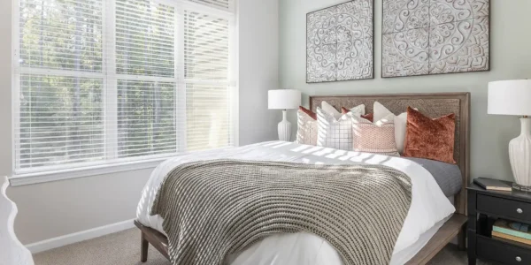 A bright, modern bedroom filled with soft daylight filtering through white horizontal blinds. The composition focuses on a plush bed layered with crisp white linens, a chunky sand-colored knit throw, and decorative pillows in rust and patterned cream. Two ornate, circular mandalas hang symmetrically on the sage-green wall above the woven headboard, flanked by elegant white lamps on dark nightstands.