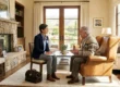 A balanced, eye-level wide shot of a female doctor in a navy blazer and an elderly man in a leather armchair, engaged in a conversation in a sun-drenched living room. Warm, natural light streams through French doors, highlighting a stone fireplace and a professional leather medical bag resting on a cream-colored rug.