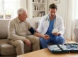 A balanced medium shot of a senior man and a male doctor seated on a beige fabric sofa. The doctor, in blue scrubs and a white coat, holds a digital blood pressure monitor while the cuff is wrapped around the patient's arm. Bright, diffused daylight fills the living room. In the foreground, an open black medical kit sits on a wooden table, revealing a stethoscope and a digital thermometer in sharp focus.