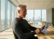 A sunlit, modern office space with floor-to-ceiling windows revealing a blurred city skyline. A mature man with grey hair, wearing a dark sweater, sits at a light wooden table, using a stylus to interact with a tablet that displays a vibrant, swirling abstract graphic of colorful lines. Warm sunlight casts long shadows across the table surface.