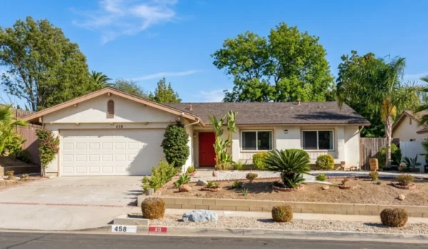 A solar-drenched, wide-angle photograph of a single-story California ranch house under a clear blue sky. Bright daylight highlights the beige stucco exterior and dark shingle roof. Clear text is visible: the white garage door has the address numbers '458' centered above it, and on the front concrete curb, the numbers '458' and '413' are painted in black. The home features a prominent red front door and a carefully manicured desert landscape with diverse plants, including cacti, barrel bushes, and large palm trees to the right. The composition centers the house, with a long concrete driveway to the left and a stone-topped retaining wall and curb in the foreground, showing the layered front yard.