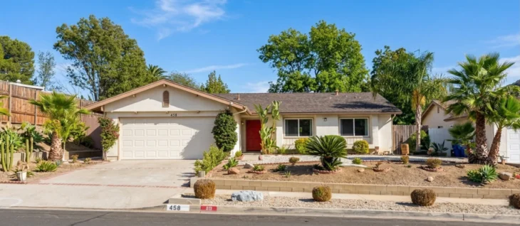caregiver-at-home-california-senior-care-home-458-s-walter-ave. A solar-drenched, wide-angle photograph of a single-story California ranch house under a clear blue sky. Bright daylight highlights the beige stucco exterior and dark shingle roof. Clear text is visible: the white garage door has the address numbers '458' centered above it, and on the front concrete curb, the numbers '458' and '413' are painted in black. The home features a prominent red front door and a carefully manicured desert landscape with diverse plants, including cacti, barrel bushes, and large palm trees to the right. The composition centers the house, with a long concrete driveway to the left and a stone-topped retaining wall and curb in the foreground, showing the layered front yard.