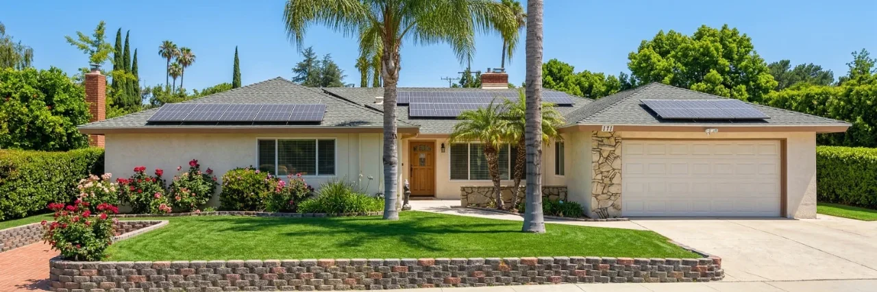 A wide-angle, bright exterior photograph of a single-story ranch-style house under a clear blue sky. The building features a tan facade, solar panels on a grey roof, and a white garage door with the number "171" clearly visible. The foreground shows a lush green lawn bordered by a tiered stone wall and blooming pink rose bushes, with two tall palm trees flanking the entrance.
