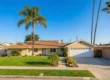 Wide-angle architectural photography of a single-story ranch house bathed in warm, golden afternoon sunlight. The composition is balanced and airy, featuring a vibrant green lawn in the foreground that leads the eye toward a beige stucco exterior and a textured brown shingled roof. Two towering palm trees frame the house against a brilliant, cloudless blue sky, creating a sense of height and serenity. The lighting casts soft, long shadows, emphasizing the flat, accessible entryway and the clean white garage door, where the house number "2676" is subtly visible. The overall aesthetic is crisp and peaceful, representing an ideal, secure environment for a caretaker for elderly at home.