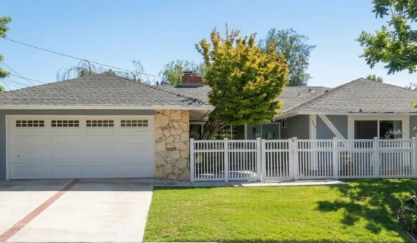 A bright, midday sun-drenched exterior photo of a single-story ranch house, seen from street level. The house is painted a muted blue-gray with a grey tiled hip roof, featuring a white panel garage door with top windows on the left. A textured wall of light-colored flagstone acts as an accent next to the garage. A distinctive turquoise front door is visible behind a long white picket fence that lines the front yard. The house numbers '5253' are clearly displayed above the entryway. Large, mature green trees frame the composition on both the left and right sides, casting defined shadows on the lush green lawn and the concrete driveway with a central brick accent stripe. A group of large agave plants is prominent in the bottom right corner. The sky is clear blue with some overhead utility lines. The overall image is clean and focused.