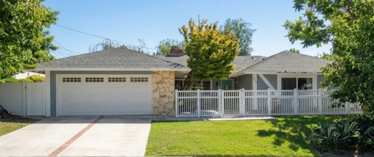 erten-home-care-thousand-oaks-for-elderly-at-home. A bright, midday sun-drenched exterior photo of a single-story ranch house, seen from street level. The house is painted a muted blue-gray with a grey tiled hip roof, featuring a white panel garage door with top windows on the left. A textured wall of light-colored flagstone acts as an accent next to the garage. A distinctive turquoise front door is visible behind a long white picket fence that lines the front yard. The house numbers '5253' are clearly displayed above the entryway. Large, mature green trees frame the composition on both the left and right sides, casting defined shadows on the lush green lawn and the concrete driveway with a central brick accent stripe. A group of large agave plants is prominent in the bottom right corner. The sky is clear blue with some overhead utility lines. The overall image is clean and focused.