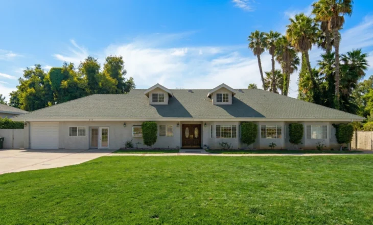 family-doctor-thousand-oaks-enduring-oaks-building A brightly lit, wide-angle exterior photograph of a sprawling, single-story light grey stucco building with a green tiled roof, designed in a residential medical office style. A broad, pristine green lawn dominates the foreground, leading to the central entrance featuring large, ornate double wooden doors. To the left, a white garage door is visible, along with a separate glass-paneled entry door for an office. The roof has two dormer windows. To the right, tall fan palm trees frame the scene against a blue sky with scattered wispy clouds. The house number '210' is clearly visible on the wall of the garage section, and another number '216' is on the central facade. The composition captures the entire building and manicured landscaping under direct sunlight.