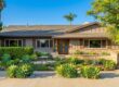A central, balanced daytime photograph of a well-appointed, contemporary single-story residence under a clear blue sky. Bright, natural sunlight highlights the textures of the warm-toned vertical siding and rough stone foundation. The entire front area features a meticulously designed xeriscape garden with large agaves and other succulents set against a field of river stones and gravel, backed by a dense, trimmed hedge. Large mature trees, including a prominent jacaranda and various cypress, frame the house on both sides. A set of glass entry doors is centered, accessed by a stone walkway and patio. In the bottom right corner, a subtle, stylized diamond-star watermark is visible.
