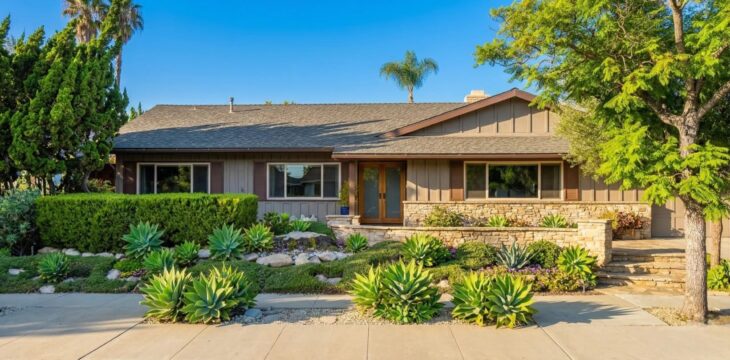 flores-compassionate-care-residence-thousand-oaks A central, balanced daytime photograph of a well-appointed, contemporary single-story residence under a clear blue sky. Bright, natural sunlight highlights the textures of the warm-toned vertical siding and rough stone foundation. The entire front area features a meticulously designed xeriscape garden with large agaves and other succulents set against a field of river stones and gravel, backed by a dense, trimmed hedge. Large mature trees, including a prominent jacaranda and various cypress, frame the house on both sides. A set of glass entry doors is centered, accessed by a stone walkway and patio. In the bottom right corner, a subtle, stylized diamond-star watermark is visible.