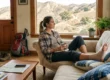 A photorealistic mid-shot of a female hiker in a plaid shirt resting on a beige sofa in a sunlit Agoura Hills home. Her right ankle is professionally wrapped in a tan elastic bandage and elevated on a blue patterned pillow. She gazes out a large window at a dry, hilly landscape. Soft, natural morning light fills the room. On her lap, a book titled "Malibu Creek State Park Trails" is visible. A red Osprey backpack and muddy hiking boots sit on the hardwood floor in the background.