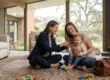 A wide, eye-level shot of a female pediatrician in a navy blue blazer sitting cross-legged on a vintage-style patterned rug in a sun-drenched, modern Thousand Oaks living room. Bright, natural daylight streams through floor-to-ceiling glass windows that reveal green, rolling hills in the background. The doctor is smiling as she uses an otoscope to examine a toddler's ear, while the child is held securely by a mother in casual attire. A classic brown leather medical bag sits in the right foreground, and colorful wooden blocks are scattered on the left, creating a balanced and reassuring clinical scene within a warm, high-end residential setting.