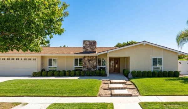 A sun-drenched, frontal photograph of a meticulously maintained suburban ranch-style home under a clear, bright blue sky. The composition is horizontally oriented, showcasing the full width of the property. Bright, direct midday sun creates crisp, defined shadows, emphasizing the textures of the beige vertical siding and the rough-hewn, stacked-stone chimney that forms the central focal point. The foreground features a concrete sidewalk and a well-manicured green lawn, separated by a row of neatly trimmed, rounded boxwood hedges. Three stepping stones lead up a concrete pathway toward the main entrance, which features double wooden doors. To the left, a large deciduous tree provides a vertical counterpoint, while a palm tree stands to the right, balancing the frame. The house number "20822" is clearly visible in a small text plaque on the stone chimney next to the door. The overall impression is one of curated, orderly, and peaceful suburban life.