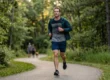 A centered, sharp-focus shot of a man in dark athletic wear running along a winding paved path in a lush forest park. The scene is bathed in warm, golden-hour light filtering through the trees, creating soft backlighting and long shadows. The composition captures the runner in mid-stride, emphasizing active lifestyle changes for cardiovascular disease prevention against a softly blurred green background.