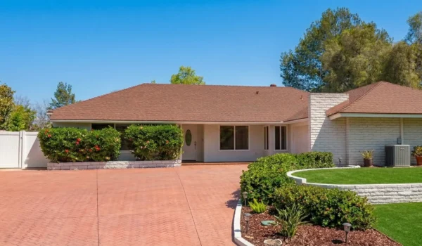 A wide-angle architectural shot of a bright, sun-drenched single-story residence featuring a clean white brick facade and a classic brown shingled roof. The composition is balanced by a prominent red-patterned driveway in the foreground and a vibrant, manicured green lawn framed by low white stone walls. Natural midday light enhances the high-contrast colors of the lush flowering bushes and the clear, cloudless blue sky, creating a serene and inviting atmosphere.