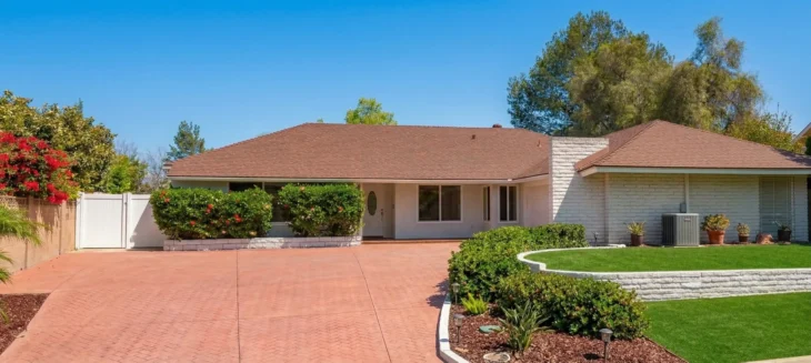 familycare-cottage-iv-elderly-inhome-care-thousand-oaks. A wide-angle architectural shot of a bright, sun-drenched single-story residence featuring a clean white brick facade and a classic brown shingled roof. The composition is balanced by a prominent red-patterned driveway in the foreground and a vibrant, manicured green lawn framed by low white stone walls. Natural midday light enhances the high-contrast colors of the lush flowering bushes and the clear, cloudless blue sky, creating a serene and inviting atmosphere.
