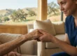 A horizontal close-up photograph bathed in soft, warm natural light from a large background window. The composition centers on a moment of connection: a compassionate middle-aged female caregiver, dressed in dark blue scrubs, leans in with a gentle smile to hold both of an elderly patient's age-spotted hands within her own. Only the patient's arms and shoulders are visible, wrapped in a knitted blanket. Through the window, a view of rolling, tree-studded hills under a clear sky creates a peaceful backdrop. The focus is sharp on their clasped hands and the caregiver's face, while the rest of the cozy room is in a soft, shallow focus