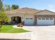 A bright, sunlit exterior photograph of a single-story home with a cream stucco and natural stone facade and a brown tiled roof. The composition captures the entire front of the house from a straight-on perspective, under a clear blue sky with light clouds. A curved concrete driveway leads to three garage doors with small windows. The entrance, under an arched portico, features a well-maintained flower bed with vibrant pink, purple, and white blooms. The house number '360' is visible above the entry. The foreground includes a lush green lawn, several small trees, and a mountain range in the distant background. The lighting is bright and even, creating crisp details and a warm, inviting feel.