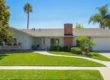 A cinematic, wide-angle architectural photograph of a single-story residential care home captured from a flat frontal perspective under a bright, cloudless blue sky. The composition is balanced, featuring a winding white concrete path that leads through a manicured, vibrant green lawn toward a central white front door. High-contrast midday sunlight creates sharp, clean shadows beneath a large leafy tree on the left and slender palm trees on the right. The building is finished with light-grey vertical siding, a prominent red brick chimney, and a clean white garage door.