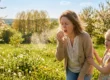 A candid medium shot in natural, warm backlight, showing a woman sneezing forcefully with closed eyes and mouth wide open, her right hand pressing a white tissue to her nose. A fine, backlit cloud of airborne particles and droplets explodes from her nose and mouth into the sunlit air. To her right, a young girl with pigtails, wearing a floral dress, has an expression of concern and is holding the woman's arm. The setting is a sun-drenched field filled with green grass, yellow dandelions, and white-flowering orchard trees, set against a distant forest line and a blue sky with soft clouds.