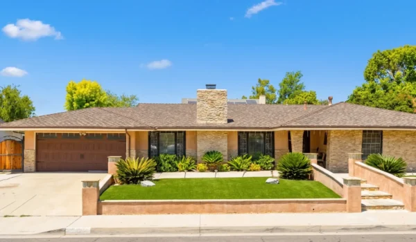 A photorealistic, wide-angle exterior view of a modern ranch-style house under a bright blue sky with a few wispy clouds. The composition is balanced horizontally, with the house centered. Direct, harsh midday sunlight illuminates the scene, casting soft shadows beneath the eaves and prominent yucca plants, creating a high-contrast and vibrant appearance. The house features light stone veneer and warm beige stucco, with a low-pitched hip roof of brown shingles. A central stone chimney is a focal point. Large windows show interior blinds. A manicured green lawn is enclosed by retaining walls. No text is visible anywhere on the object or in the scene.