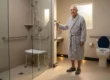 A wide-angle architectural composition bathed in soft, diffused morning sunlight filtering through a frosted window on the right. The warm, cinematic lighting casts gentle, realistic shadows across matte beige floor tiles, complemented by subtle amber motion-sensor LED lighting along the baseboards. Positioned slightly right of center to balance the frame, a dignified elderly man in a textured light gray bathrobe and non-slip slippers maintains a natural, stable posture. His right hand is securely gripping a vertically mounted, brushed nickel wall support. To the left, a glass-enclosed zero-threshold shower stall contains a white, medical-grade shower bench and horizontal metal supports. To the right, a white comfort-height toilet is visually anchored by a high-contrast dark blue lid. The overall aesthetic is highly detailed, premium, and structurally realistic, with no visible text or branding on any objects or surfaces.