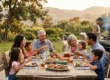 A cinematic, wide-angle composition illuminated by warm golden-hour sunlight casting a soft glow across a wooden outdoor dining table. The image features a layered depth of field, keeping the seated family and fresh food in sharp focus, while a figure operating a BBQ grill in the midground and the rolling mountainous background fade into a softly blurred bokeh.