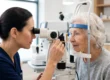 A profile photograph of an eye examination, with an optometrist and an elderly patient on opposite sides of a complex white and metal slit-lamp instrument. The intricate optical device is the central, sharply focused element, standing between the two women. The scene is evenly lit by cool, clean clinical overhead lighting from the left and top, highlighting textures and form. The optometrist on the left, with dark hair tied back, peers into an eyepiece. The patient on the right, with grey hair and a blue headband, gazes into the corresponding viewer, her head supported. Their faces frame the machine. The clinic background is a soft blur of shelves and bottles.