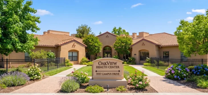 A wide-angle, head-on photograph capturing the symmetrical exterior of a single-story Spanish-style building under a bright blue sky with scattered clouds, as seen in oakview-health-center-manicured-exterior.jpg. A brick paver pathway in the foreground leads the eye to a large monument sign in the center, flanked by dark metal fencing and manicured beds of purple lavender, pink-tipped roses, and large blue-purple hydrangeas. The central sign, in a serif font, clearly states from top to bottom: 'OAKVIEW', 'HEALTH CENTER', a line separator, and '3557 CAMPUS DRIVE'. The building behind, with beige stucco and terracotta tile roofs, features multiple arched entries and large windows, with potted palms near the door. Bright daylight illuminates the scene and defines sharp shadows, emphasizing the textures of the roof tiles, stucco, and pavers.