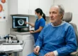 A candid, brightly lit photograph taken from a slightly low angle in a clinical pre-operative eye surgery room. On the right, an elderly Caucasian man with grey, thinning hair and a beard, dressed in a blue surgical gown, is seated in a tan medical chair, looking off-camera to the left. In the middle ground on the left, a female medical assistant with her dark hair pulled back and wearing a blue mask and scrubs is standing, pointing at a circular tomographic eye scan image on a computer monitor. Further to the far left, a rollable metal medical cart holds various items, including handheld instruments, bottles of eye drops, and a paper document. A detailed anatomical diagram of the human eye hangs on the wall behind the cart. The overall lighting is cold fluorescent, and the composition is balanced.