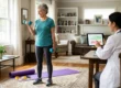 A photograph of a grey-haired woman in teal sportswear, actively holding blue dumbbells, smiling towards a seated consultant in a white medical coat in a bright, sun-drenched living room setting. The consultant, seen from behind at a table, is pointing at a graph on a tablet screen, which shows green, yellow, and red zones. The woman is standing on a purple yoga mat, with a yellow resistance band and large windows in the detailed background. The natural light emphasizes the comfortable, home environment.