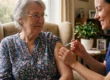 A medium close-up photograph capturing a calm interior scene bathed in warm natural light. Diffused sunlight from a large window on the left illuminates a senior woman with grey hair and glasses, seated in a textured, beige armchair. She is wearing a blue floral patterned blouse and is looking calmly towards a care worker to her right. The care worker, wearing a blue scrub top and with a gentle smile, is in profile. Her hands are in sharp focus, precisely administering a clear syringe with a needle into the older woman's upper left arm. The background is a softly blurred, detailed living room with books on shelves and framed photographs. The composition is intimate, focusing on the careful act of injection. No text is visible on the subjects or main objects.