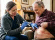 A warmly lit, medium-shot photograph composed with two main figures in a cozy indoor home setting. Soft, natural daylight streams in from the left side, gently highlighting the side profile of a female healthcare worker on the left and the face and torso of an older man seated on a textured sofa to the right. The composition focuses intimately on the interaction: the healthcare worker, wearing bright blue nitrile gloves, leans in with a metallic penlight, casting a distinct bright beam onto the man's exposed lower abdomen. The skin reveals a striking, textured band of vivid red blisters. The background features a soft depth of field, blurring warm wooden furniture, books, and a glowing table lamp, which creates an intimate and caring atmosphere. There is no visible text in the image.