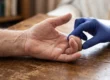 A close-up, horizontal composition with a shallow depth of field, illuminated by soft, diffused natural daylight. Entering from the left, an older, heavily textured hand rests palm-up on a weathered wooden table, with its ring and pinky fingers distinctly bent inward. Entering from the upper right, a hand wearing a bright blue nitrile glove sharply contrasts with the warm tones of the skin and wood, its fingers delicately touching the bent digits. The background is a soft, out-of-focus blur of warm, muted interior elements.
