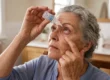 Soft, diffused natural window light from the left illuminates a close-up, slightly low-angle composition. The subject, an older woman, is sharply in focus in the center foreground, tilting her head back while gently pulling down her lower eyelid. A shallow depth of field softly blurs the warm-toned, neutral bathroom setting in the background. She is holding a small, clear plastic dropper bottle over her eye, which features a label with the legible text "DRY EYE RELIEF" and "10ml".